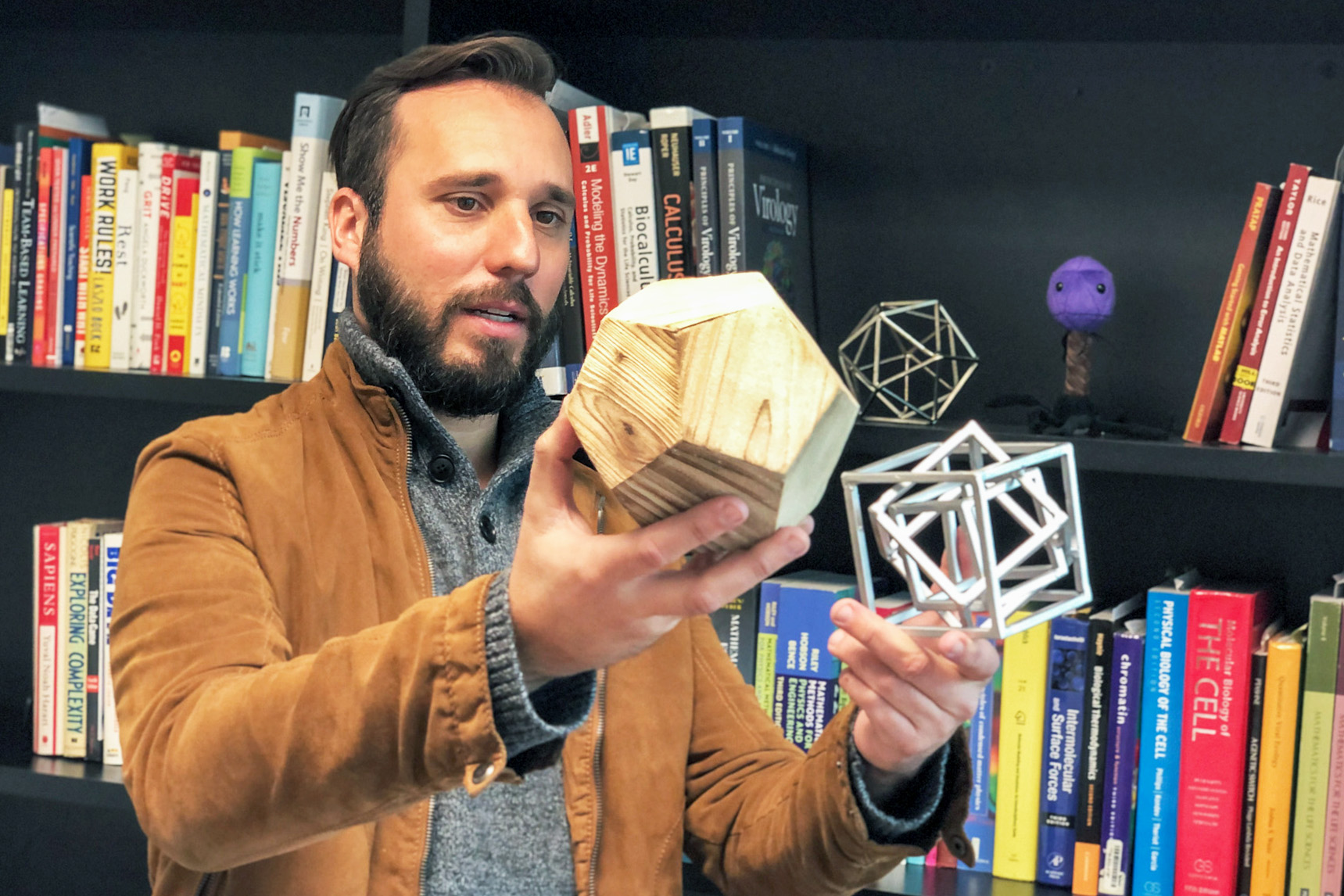 A man holds up a wooden icosahedron and a metal cube within a cube within a cube as models of virus capsids.