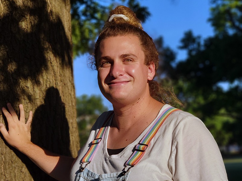 Brienne, with curly blond hair in a half up bun, wears a white t-shirt and denim overalls with rainbow suspenders. They are outside, smiling, while touching a tree trunk.